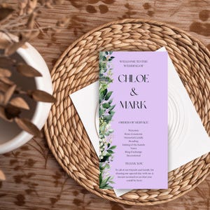 May include: A wedding program with a lavender background and floral accents. The text reads "Welcome to the Wedding of Chloe & Mark" and lists the order of service. The program is on a white card, placed on a woven placemat.