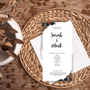 以下が含まれることがあります： 「Sarah & Mark」の名前がエレガントな筆記体で書かれた結婚式のプログラム。白いカードには、ネイビー、オレンジ、ティール色の花のアクセントが施されています。プログラムには、サービスの順序と感謝のメッセージが含まれており、織りのプレースマットの上に置かれています。