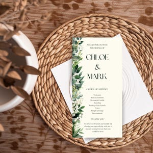 May include: A wedding program featuring the names "Chloe & Mark" in black. The program is adorned with green leaves and white flowers. The text includes "Order of Service" and a "Thank You" message. It rests on a woven placemat.