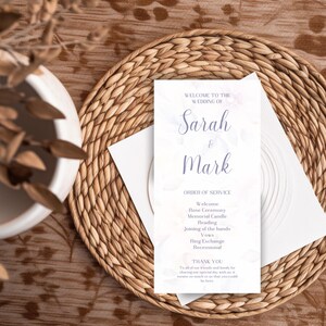 Pode incluir: Um programa de casamento com os nomes "Sarah & Mark" em escrita elegante. O programa inclui a ordem da cerimónia, impresso num cartão branco com um design floral. O cartão repousa sobre um descanso de mesa tecido.