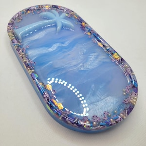 May include: An oval-shaped resin tray with a light blue ocean design and a light blue palm tree. The tray's border is decorated with purple flowers, iridescent glitter, and small white beads. The tray is approximately 6 inches long and 3 inches wide.