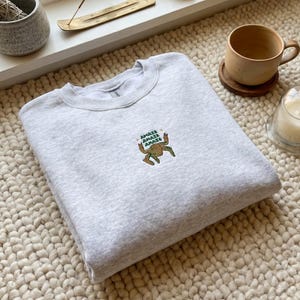 Peut inclure: Un sweat-shirt gris clair à col rond, orné d'une broderie représentant une grenouille avec le mot "Amaze". Le sweat-shirt est plié sur un tapis texturé près d'une fenêtre.