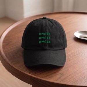May include: A black baseball cap featuring the word AMAZE embroidered in green thread three times, stacked vertically on the front panel. The cap has a structured crown, a curved brim, and a button at the top center of the fabric dome.