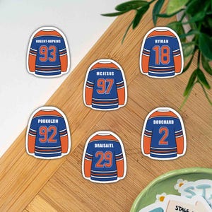 Könnte beinhalten: Sechs Hockey-Trikot-Aufkleber in Blau und Orange. Jeder Aufkleber zeigt den Namen und die Nummer eines Spielers, darunter Nugent-Hopkins, McJesus, Hyman, Podkolzin, Draisaitl und Bouchard. Sie liegen auf einer Holzoberfläche.