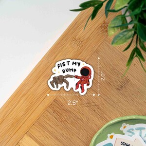 May include: A die-cut sticker featuring a brown creature and a red astronaut fist-bumping, with the text FIST MY BUMP in black letters. The sticker measures 2.5 inches wide by 2.0 inches high, as indicated by the visible measurement markings.
