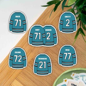 Könnte beinhalten: Eine Sammlung von tealfarbenen Hockey-Trikot-Aufklebern mit weißen und schwarzen Streifen. Jeder Aufkleber zeigt einen anderen Namen und eine andere Nummer, darunter "MACK 71", "SMITTY 2", "EKLUND 72", "MISA 77" und "WENNBERG 21". Der Aufkleber "BABY SHARKS 71-2" ist ebenfalls enthalten.
