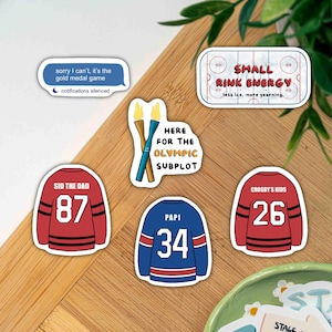 May include: A collection of hockey-themed stickers. Designs include hockey jerseys with numbers, a hockey rink graphic, a text bubble with the phrase "sorry I can't, it's the gold medal game", and Olympic torch graphics.