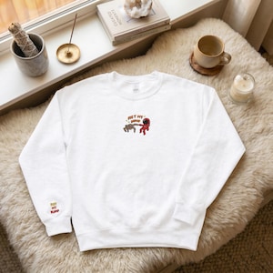 May include: White sweatshirt with embroidered design of two astronauts fist bumping. The text "Fist My Bump" is embroidered on the sleeve and above the astronauts. The sweatshirt is made of a soft material.