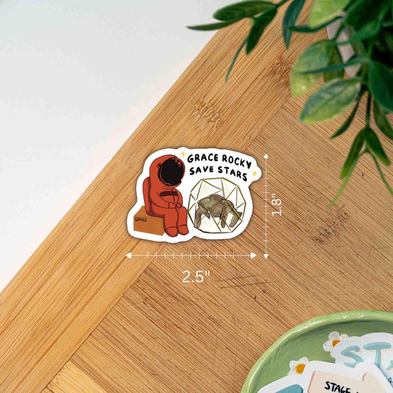 May include: A die-cut sticker featuring a red astronaut sitting on a box labeled Grace, next to a geometric cage containing a turtle. The text reads GRACE ROCKY SAVE STARS. A white ruler overlay indicates dimensions of 2.5 inches by 1.8 inches.