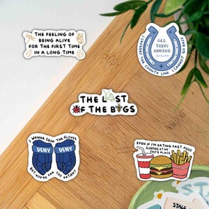 Peut inclure: Cinq autocollants en vinyle avec des motifs variés : un texte sur le sentiment d'être vivant, un fer à cheval bleu avec du texte, un autocollant THE LAST OF THE BUGS avec des insectes, des gants de hockey bleus et un repas fast-food.