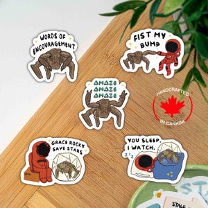 May include: Five vinyl stickers featuring brown, multi-legged alien creatures and red-suited astronauts. Text includes "WORDS OF ENCOURAGEMENT," "FIST MY BUMP," "AMAZE," "GRACE ROCKY SAVE STARS," and "YOU SLEEP. I WATCH." Designs include stars and geometric shapes.