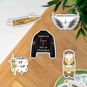May include: A collection of five stickers featuring a gold bullet with the text Memento Mori, a pair of grey wings with the text Umbra Mortis, a black jacket with the text Through Love All Is Possible, a star graphic with Light It Up, and an otter.