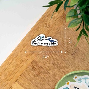 Pu&ograve; includere: Un adesivo bianco con la scritta "Don't marry him" e un disegno di onda blu. L'adesivo &egrave; lungo 6,1 cm e alto 2,8 cm. L'adesivo &egrave; su una superficie di legno.