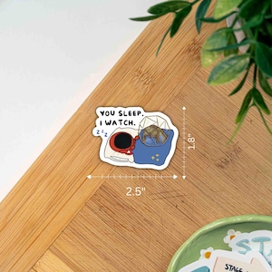 May include: A die-cut sticker featuring a red crab in bed with a white pillow and blue blanket, next to a geometric cage containing a small creature. The text reads "YOU SLEEP. I WATCH." with "Zzz" symbols. A ruler indicates dimensions of 2.5" by 1.8".