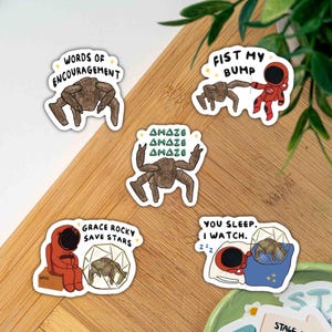 Op de afbeelding: Een verzameling eigenzinnige stickers met een bruin insectachtig wezen en een astronaut. De stickers tonen tekst als "Words of Encouragement", "Fist My Bump", "Amaze", "Grace Rocky Save Stars" en "You Sleep. I Watch."