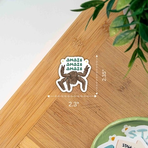 May include: A die-cut sticker featuring a brown, multi-legged creature with the text "AMAZE" repeated three times in teal above it. The sticker measures 2.3 inches in width and 2.35 inches in height as indicated by the visible measurement lines.