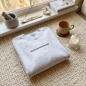 Könnte beinhalten: Ein hellgraues Sweatshirt mit Rundhalsausschnitt und dem schwarzen gestickten Text NULLUM GRATUITUM PRANDIUM mittig auf der Brust. Das Kleidungsstück besteht aus meliertem Stoff und ist ordentlich auf einer cremefarbenen Oberfläche gefaltet.