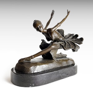 Könnte beinhalten: Eine bronzefarbene Skulptur einer tanzenden Person in dynamischer Pose mit nach oben gestreckten Armen und einem nach vorne ausgestreckten Bein. Die Figur trägt ein Tutu mit geschichteten, blütenblattartigen Falten auf einem ovalen Steinsockel.
