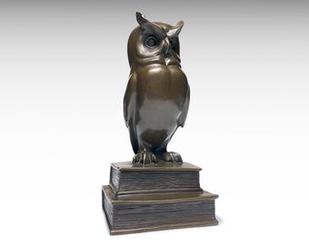 May include: A dark bronze-colored sculpture of an owl standing upright on top of two stacked rectangular books. The owl has prominent ear tufts and detailed feathers, while the books feature textured edges representing pages in a solid metal finish.