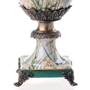 Op de afbeelding: Een witte keramische vaas met een groen en gouden bloemenmotief. De vaas heeft een vierkante basis met sierlijke gouden voetjes en een rond lichaam met een gouden rand.