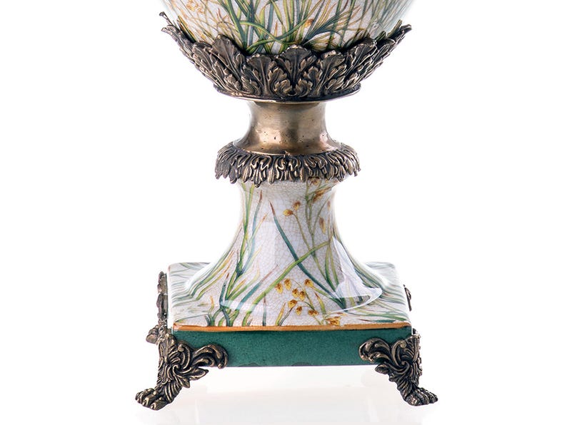 Op de afbeelding: Een witte keramische vaas met een groen en gouden bloemenmotief. De vaas heeft een vierkante basis met sierlijke gouden voetjes en een rond lichaam met een gouden rand.