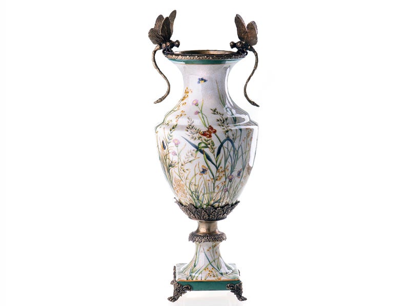 Op de afbeelding: Een witte keramische vaas met een bloemenmotief en gouden accenten. De vaas heeft twee libellenhandvatten en staat op een vierkante voet met gouden poten.