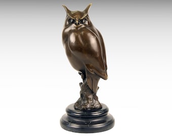 May include: A bronze-toned owl sculpture perched on a textured branch base. The owl features pointed ear tufts, large dark eyes, and smooth plumage. The entire figure is mounted on a circular, polished black base, creating a solid, weighted appearance.