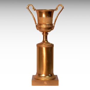 Puede incluir: Un trofeo dorado con un diseño clásico de urna y dos asas. El trofeo se asienta sobre una base cuadrada.
