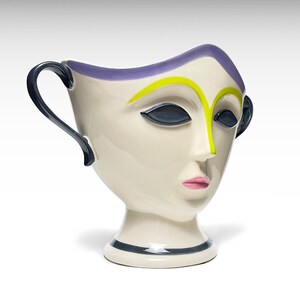 May include: A ceramic pitcher shaped like a human face with a cream-coloured base, dark blue handles, a purple rim, yellow eyebrows, dark blue almond-shaped eyes, and pink lips. The vessel features a pedestal base with a dark blue horizontal stripe.