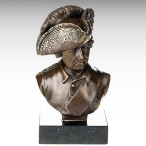 May include: Bronze bust sculpture of a man wearing a military uniform and a feathered hat. The bust is on a black marble base.