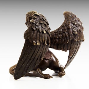 Puede incluir: Escultura de bronce de una figura alada con cabeza humana y cuerpo de le&oacute;n. La figura est&aacute; arrodillada con las alas extendidas detr&aacute;s de ella. La escultura est&aacute; firmada en la parte posterior.