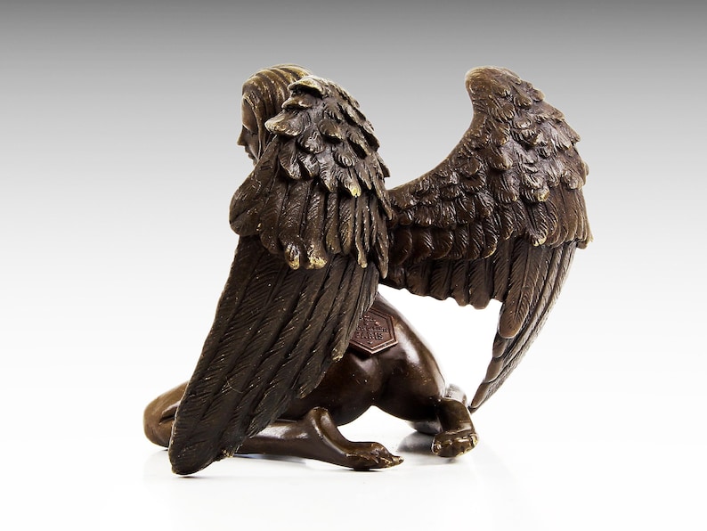 Puede incluir: Escultura de bronce de una figura alada con cabeza humana y cuerpo de le&oacute;n. La figura est&aacute; arrodillada con las alas extendidas detr&aacute;s de ella. La escultura est&aacute; firmada en la parte posterior.