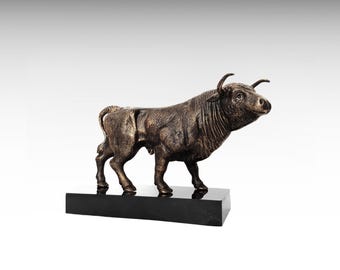 May include: A bronze-toned bull figurine with a textured, muscular body and curved horns, standing in a forward-facing pose. The sculpture is mounted on a rectangular, polished black base that provides a stable foundation for the piece.
