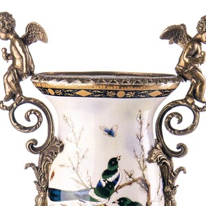 May include: A white ceramic vase with a painted bird and floral design. The vase has ornate bronze handles and a gold and black rim. Two bronze cherub figures are perched on the handles. The vase is a decorative item.