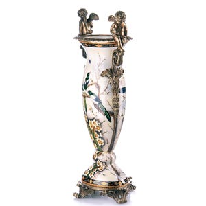 May include: An ornate vase with a white and marble effect finish, decorated with a painted bird and floral design. Two cherub figures sit on top. The base and accents are gold-toned.