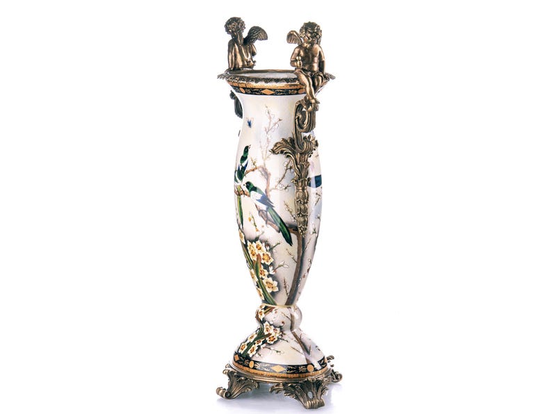 May include: An ornate vase with a white and marble effect finish, decorated with a painted bird and floral design. Two cherub figures sit on top. The base and accents are gold-toned.