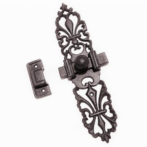 May include: A dark gray cast iron door latch featuring an elongated backplate with intricate openwork scroll patterns and a fleur-de-lis motif. The hardware includes a sliding bolt mechanism with a round knob and a separate rectangular strike plate.