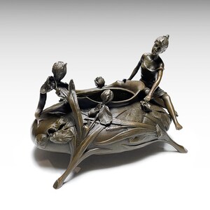 Pode incluir: Um centro de mesa de metal em tom de bronze com uma figura esculpida sentada na borda. O recipiente é decorado com motivos florais em relevo, incluindo flores de íris e folhas longas, e assenta sobre três pernas curvas e afiladas.