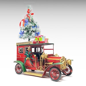 Puede incluir: Un modelo de coche rojo vintage con detalles verdes y dorados. Un pequeño árbol de Navidad y regalos envueltos están en la parte superior. El coche tiene ruedas rojas y un lazo verde decorativo.