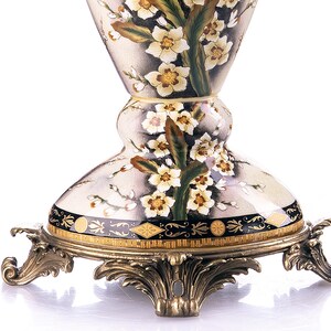 May include: A decorative vase with a floral design. The vase has a cream background with painted white and yellow flowers and green leaves. The base is a gold-coloured metal with ornate detailing.