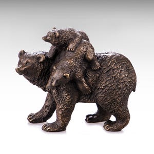 Puede incluir: Una escultura de tono bronce que representa a un oso adulto grande de pie de perfil con dos oseznos más pequeños posicionados sobre su espalda. La superficie de la escultura presenta un acabado texturizado similar al pelaje con reflejos marrones.
