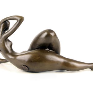May include: Bronze sculpture of a nude figure lying on its side. The figure is posed with one arm raised above its head and the other arm resting on its hip. The sculpture is made in a realistic style and has a smooth, polished finish.