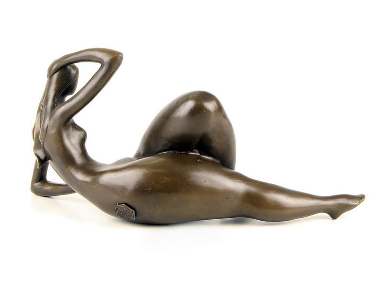 May include: Bronze sculpture of a nude figure lying on its side. The figure is posed with one arm raised above its head and the other arm resting on its hip. The sculpture is made in a realistic style and has a smooth, polished finish.