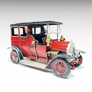 Puede incluir: Un modelo de coche antiguo rojo con guardabarros y techo negros. El modelo presenta faros y una bocina dorados, con ruedas rojas. El coche tiene un interior detallado y está sobre un fondo blanco.