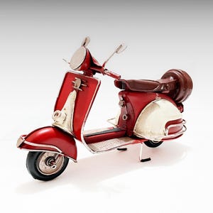 Puede incluir: Un modelo de scooter rojo y crema de estilo vintage. El scooter presenta un diseño clásico con carrocería roja, detalles en crema y asiento marrón. El modelo tiene un diseño detallado con portaequipajes y espejos.