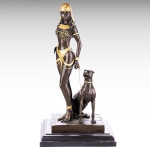 May include: Bronze sculpture of a woman in a gold and black costume holding a leash with a dog. The woman is wearing a headdress and the dog is sitting.