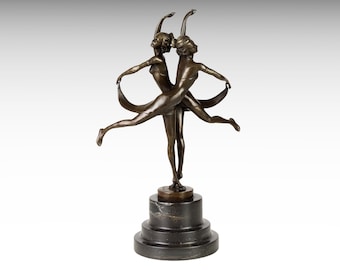 Op de afbeelding: Bronzen sculptuur van twee danseressen in een sierlijke pose, armen geheven en benen uitgestrekt. De danseressen zijn verbonden door een vloeiend lint. De sculptuur is gemonteerd op een donkere, gelaagde marmeren voet.