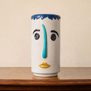 May include: A cylindrical ceramic vase with a white base featuring a stylised face. The design includes a textured dark blue rim, dark blue eyes and eyebrows, a prominent turquoise nose, and yellow lips. The surface is smooth and glossy.