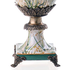 Op de afbeelding: Een witte keramische vaas met een groen en gouden bloemenmotief. De vaas heeft een vierkante basis met sierlijke gouden voetjes en een rond lichaam met een gouden rand.