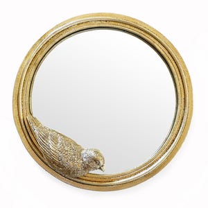 Op de afbeelding: Ronde decoratieve spiegel met een goudkleurige lijst. Een gedetailleerde, getextureerde vogelsculptuur is aan de lijst bevestigd. Het oppervlak van de spiegel is reflecterend en het ontwerp is elegant en sierlijk.